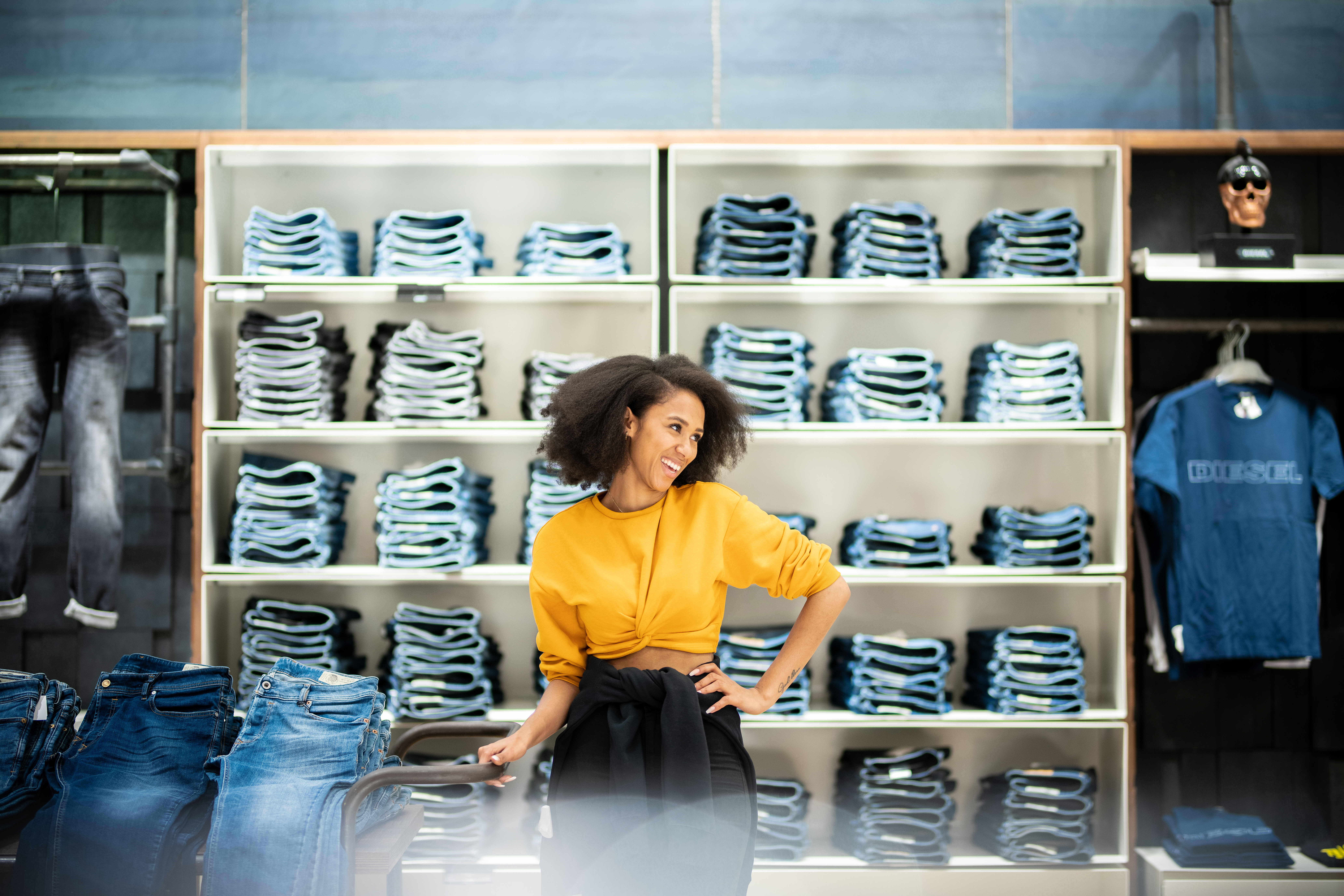 Junge Frau mit schwarzen Locken und gelbem cropped Pullover steht im Verkaufshaus vor einer Wand mit Jeans und lächelt zur Seite.
