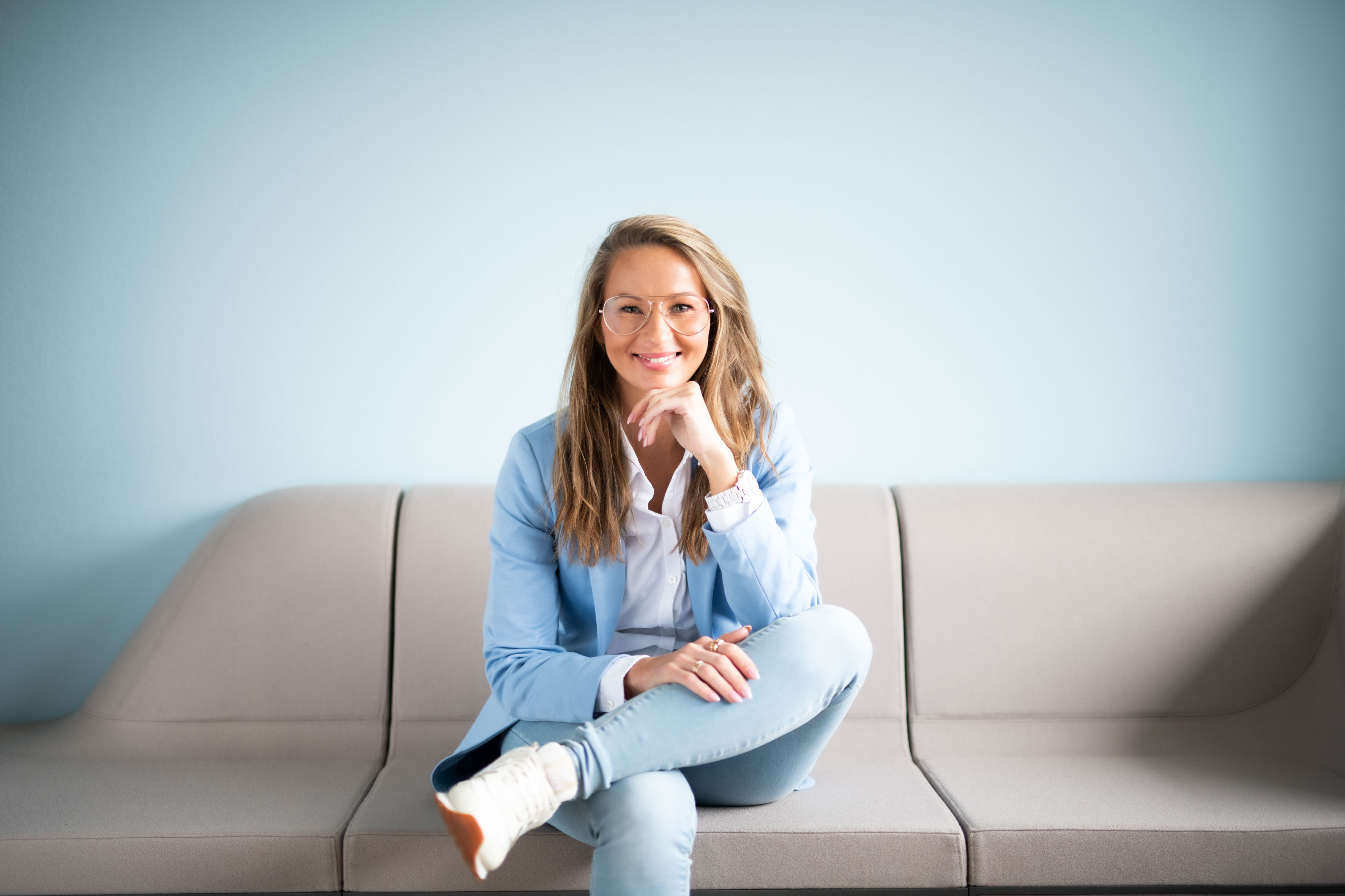 Eine blonde Frau mittleren Alters mit Brille und blauem Blazer sitzt auf einem Sofa und lächelt.